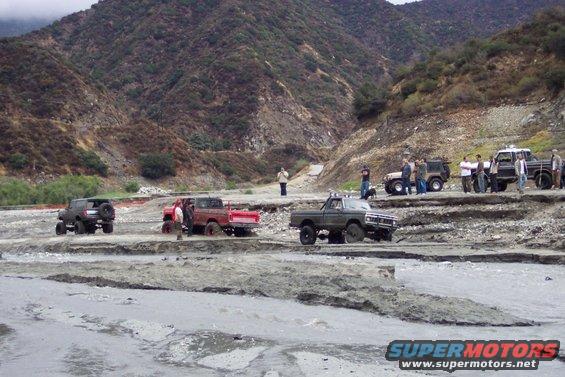 Several off-road trucks and SUVs navigate a muddy, rocky terrain with steep hills in the background