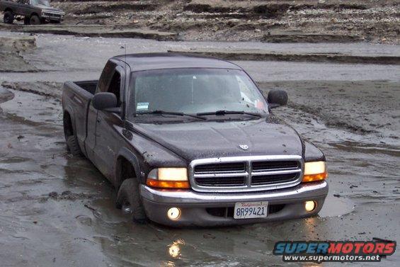 Dark pickup truck stuck in mud, front view. Another older truck is visible in the background