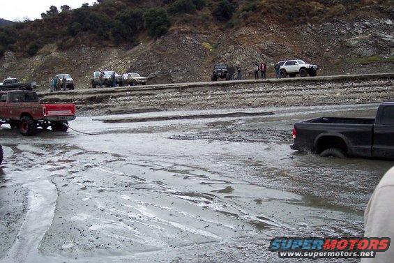 Several trucks and SUVs are stuck in a muddy, wet terrain with a rocky hillside in the background