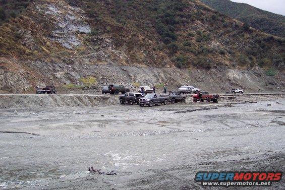 Multiple trucks and SUVs parked in a rocky, dry riverbed with a mountain in the background