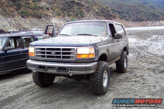 Ford Bronco parked on muddy ground next to a dark SUV. Hills in background