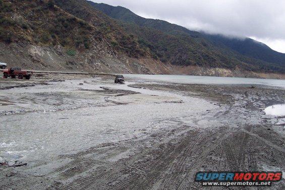 Dark SUV driving through shallow water and mud near a lake, with a red pickup truck and white vehicle parked on the shore