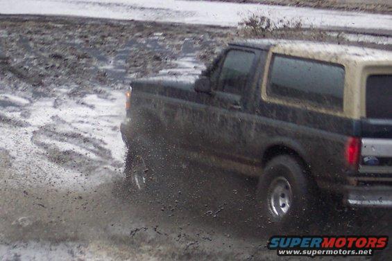 Dark Ford Bronco with tan top driving through mud, rear quarter view