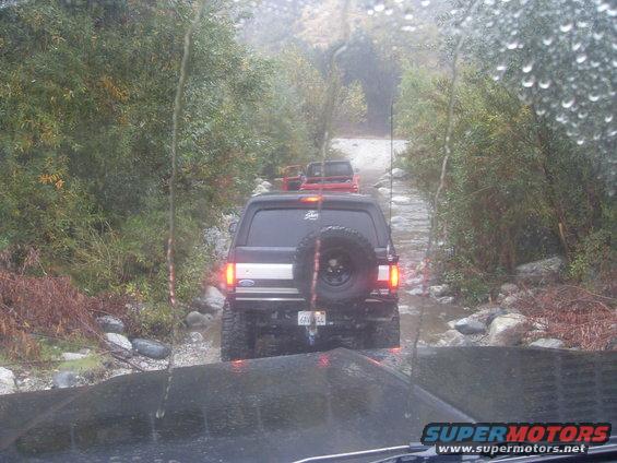 Several SUVs driving on a rocky, wet trail. The lead vehicle is a black Ford Bronco with a spare tire on the back