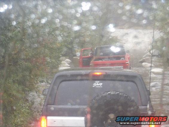 Dark SUV rear view with spare tire, followed by red pickup truck with open hood