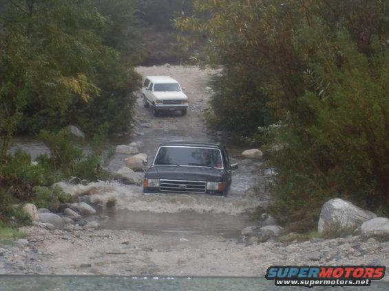 Dark SUV driving through water, white SUV behind it on a dirt path