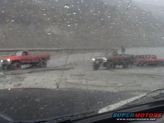 Three red pickup trucks and a red SUV drive on a wet, muddy road