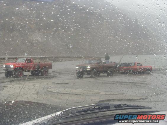 Three lifted trucks driving through mud; one red pickup, one brown pickup, and one red SUV
