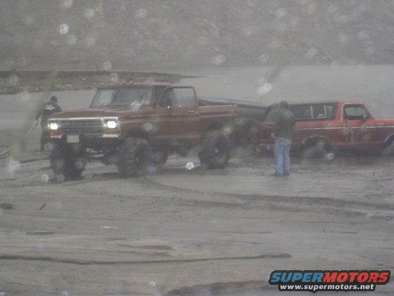 Muddy brown truck and red truck in a muddy area. One person stands by the red truck