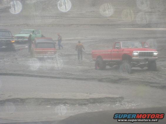 Red pickup truck with lifted suspension driving in mud, other vehicles visible