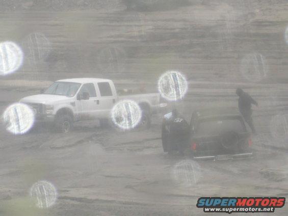 Black SUV with spare tire on rear, in a muddy scene with a white pickup truck