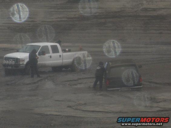 White pickup truck and black SUV parked in a muddy, off-road area with two people standing nearby