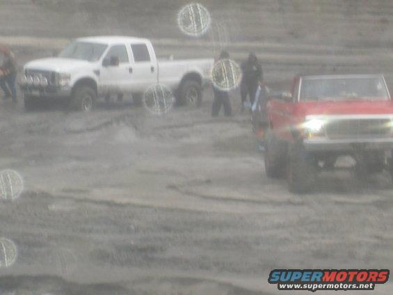 Two pickup trucks, one white crew cab and one red, are in a muddy area with people around them