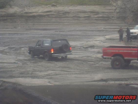 Black 1978 Ford Bronco with spare tire on rear, being towed by red pickup truck in sandy terrain