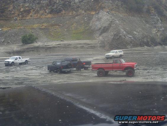 Several trucks and SUVs are off-roading in a muddy, rocky area. A red pickup truck is prominent