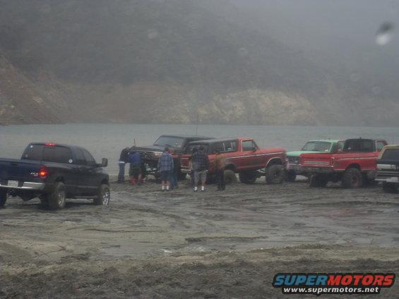 Group of trucks and SUVs gathered on a muddy shore near water, with a 1979 Ford Bronco among them