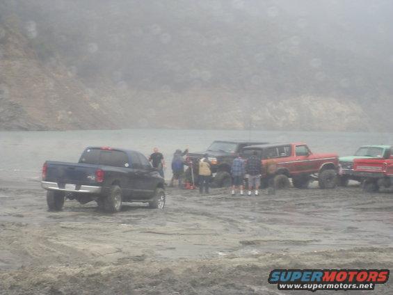 Several trucks and SUVs gathered on a sandy area near water, with people standing around them