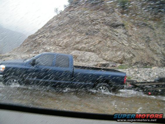 Blue pickup truck towing a trailer through flooded road with rocky hillside in background