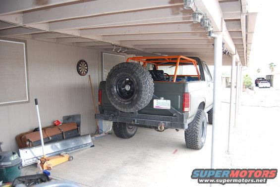 Rear view of a dark green 1982 Ford Bronco with an orange roll cage and a spare tire mounted on the back. It's parked under a carport