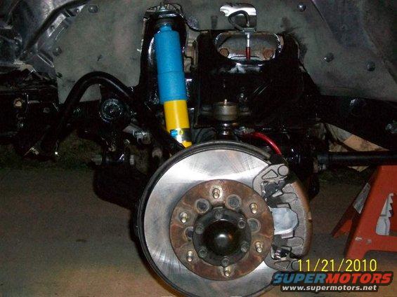 Close-up of a vehicle's front suspension with a blue and yellow Bilstein shock absorber and a disc brake assembly. A red jack stand is visible on the right. Watermark reads 11/21/2010 SUPERMOTORS.NET