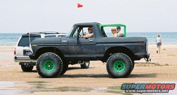 Lifted black Ford Bronco with green wheels on a beach, people inside. Another SUV is parked behind it
