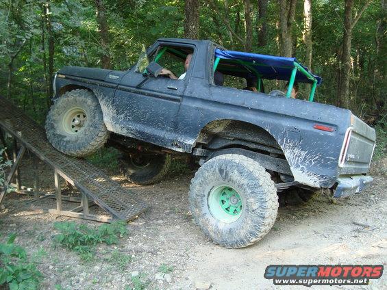 Black off-road truck with green roll cage going up a metal ramp in woods. It has large tires and a blue soft top