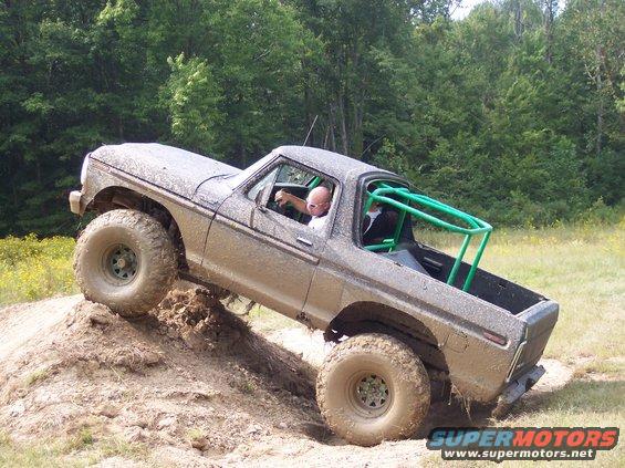 Mud-covered 1978 Ford Bronco with green roll cage climbing a dirt mound