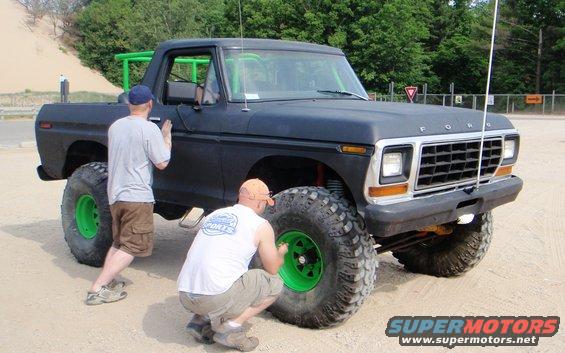 Black 1979 Ford Bronco with green roll cage and bright green wheels, front 3/4 view, two men working on it