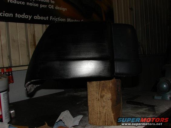 Black plastic automotive part, possibly a fender flare or bumper section, resting on a wooden block