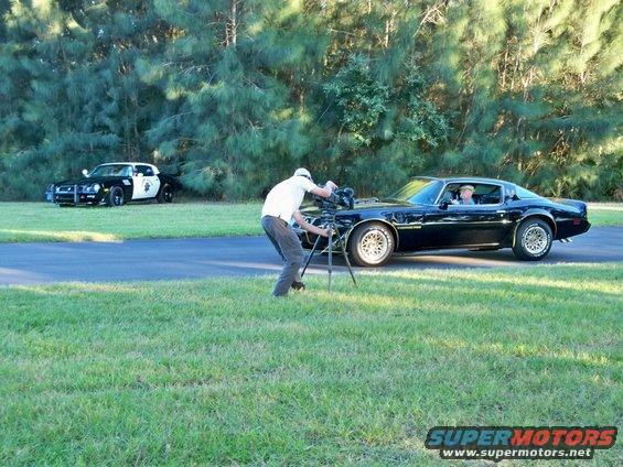 Guy filming a black 1977 Pontiac Trans Am with a police car in the background