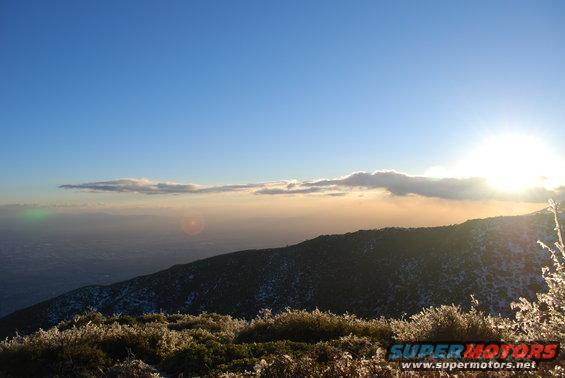 Mountain landscape with sun rising over clouds, frost on foreground plants, and a "SUPER MOTORS" watermark