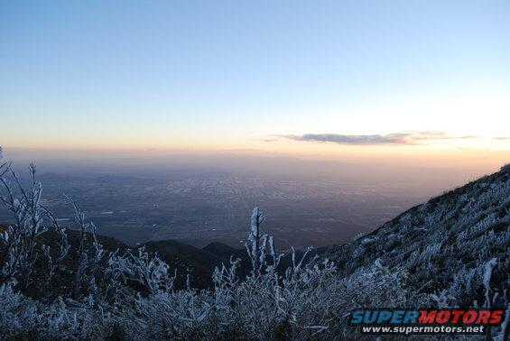 Mountain landscape with frost-covered branches in the foreground and a hazy city visible in the distance at sunrise
