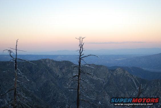 Mountain range with dead trees in the foreground at sunset, sky fades from yellow to pink to blue