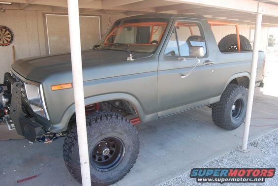 Olive green Ford Bronco with orange roll cage and spare tire in the rear, parked under a carport