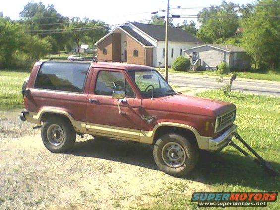 1986 Ford Bronco II parked on gravel with a flat front tire, red paint, tan lower trim