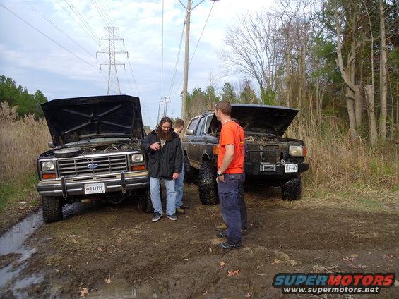 Two Ford Broncos with hoods open stuck in mud, people standing around them