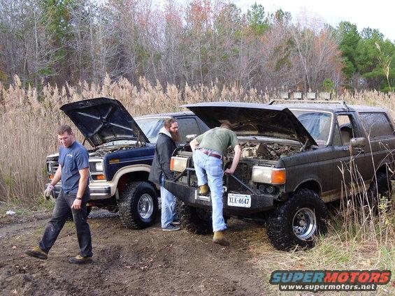 Two off-road vehicles with hoods up, one a dark Bronco, the other a blue pickup. People are working on them in a muddy, grassy area