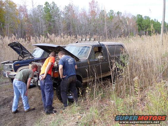 Dark brown Ford Bronco stuck in mud with hood open, three men examining engine