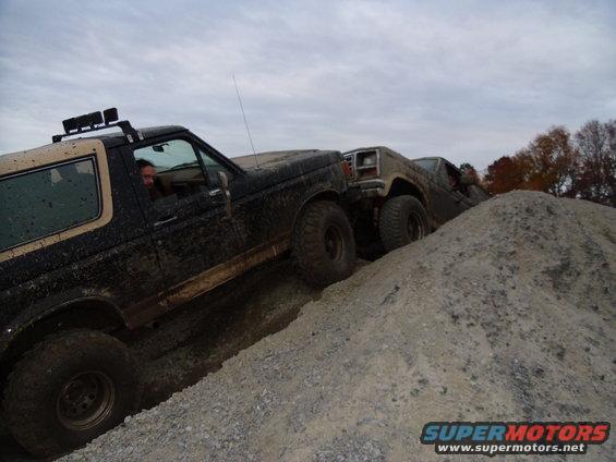 Two off-road SUVs climbing a steep dirt mound. The lead vehicle is black with a tan top, and the second is tan