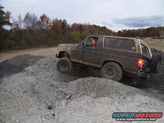Muddy brown 1985 Ford Bronco driving over gravel, rear-mounted spare tire visible