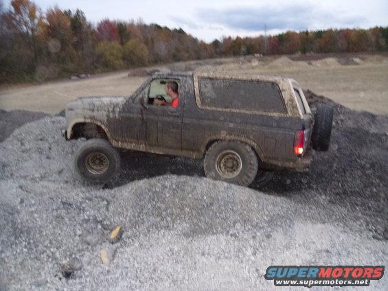 Muddy 1980s Ford Bronco climbing over gravel piles. Driver visible inside