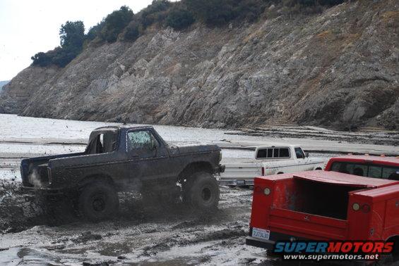 Muddy trucks driving through a wet, rocky area with a large hill in the background