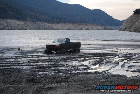 Dark-colored Ford F-Series truck with large tires stuck in mud near a body of water