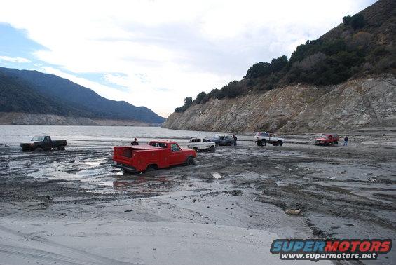 Several trucks stuck in mud near a lake with a rocky hillside