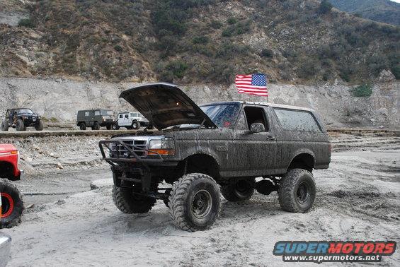 1996 Ford Bronco, lifted and covered in mud, hood open, with an American flag on the roof. Other off-road vehicles visible in the background