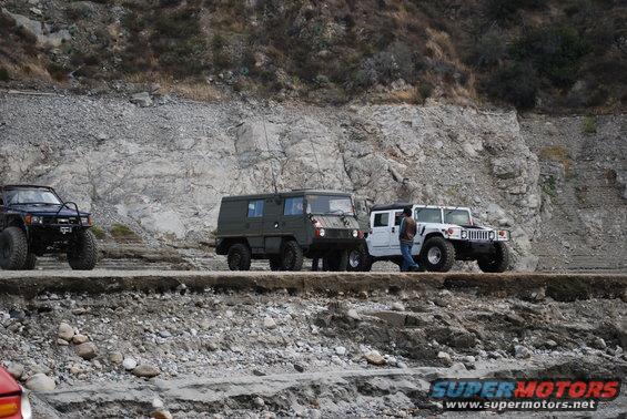 Four off-road vehicles, including a dark SUV, a green military-style van, and two white Hummers, parked on a dirt road next to a rocky hillside