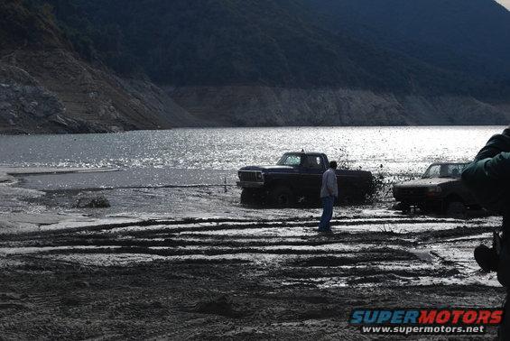 Dark purple truck stuck in mud, facing left, with a person standing nearby. Another truck is visible to the right