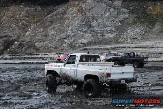 Several lifted trucks driving through mud, one white pickup in foreground