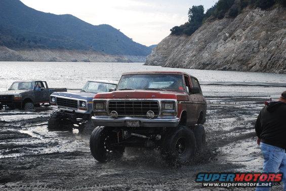 Red lifted Ford Bronco driving through mud near a lake