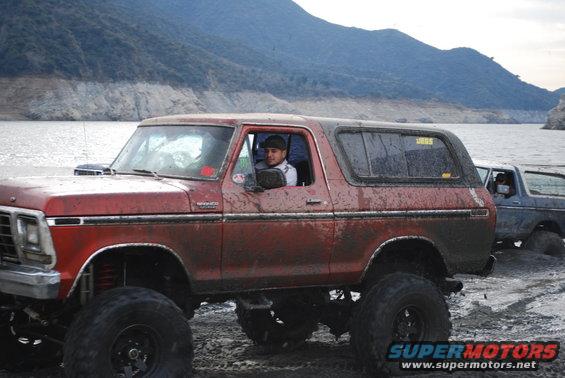 Red Ford Bronco with lifted suspension and large tires, covered in mud, parked next to another vehicle by a body of water